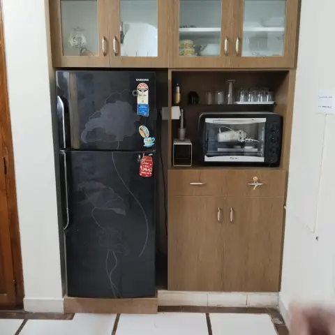 Kitchen tall-unit alcove housing a black refrigerator beside an open shelf with a toaster oven and glass-front crockery cabinets above.