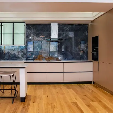 Kitchen with a dramatic dark blue-grey marble slab backsplash behind a chimney hood, taupe matte cabinetry, wood flooring and a breakfast bar with stools