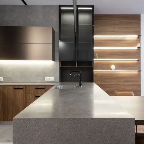 Kitchen with a grey terrazzo waterfall island, walnut cabinetry, and a backlit walnut shelving display wall, opening onto a dining table.
