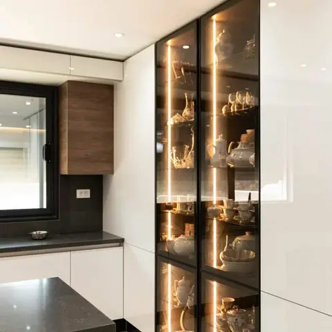 Kitchen with glossy white cabinets and a black stone countertop, anchored by a floor-to-ceiling glass-front crockery display cabinet lit with vertical LED