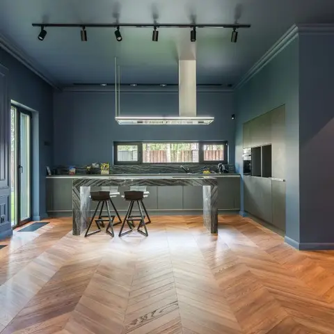 Kitchen with navy-blue walls, a marble waterfall island with bar stools, chevron-pattern wood flooring, and full-height glass doors opening to a garden.