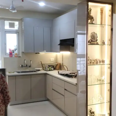 L-shaped beige gloss modular kitchen beside a tall backlit glass display cabinet showcasing trophies, crystal glassware and crockery.