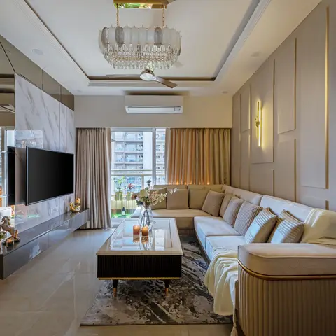 L-shaped beige living room with a mirrored-and-marble TV feature wall, brass wall sconces, a crystal chandelier over a cove-lit tray ceiling