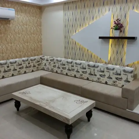 L-shaped beige sofa with Paris-print bolster backs sits under a backlit grey diamond panel on wavy-patterned wallpaper