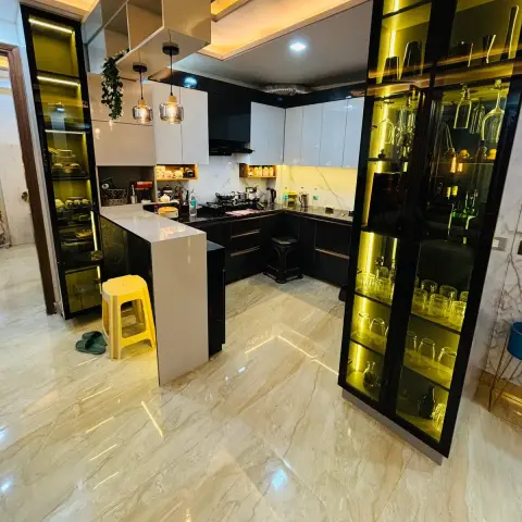 L-shaped black-and-white glossy modular kitchen with backlit glass crockery and bar units flanking a marble breakfast counter, on cream marble flooring.