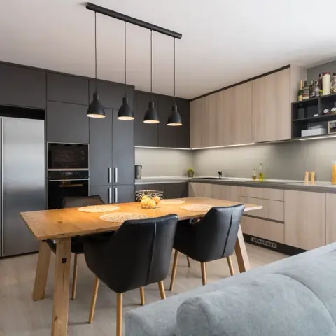 L-shaped charcoal-and-wood kitchen with a wooden dining table under black pendant lights, opening onto a grey sofa in the foreground.