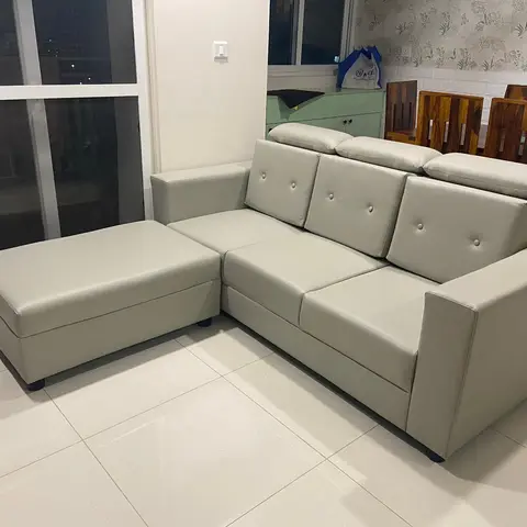 L-shaped grey sofa with a matching ottoman in a living-dining space with floral wallpaper and a sheesham wood dining table with carved chairs in the background.