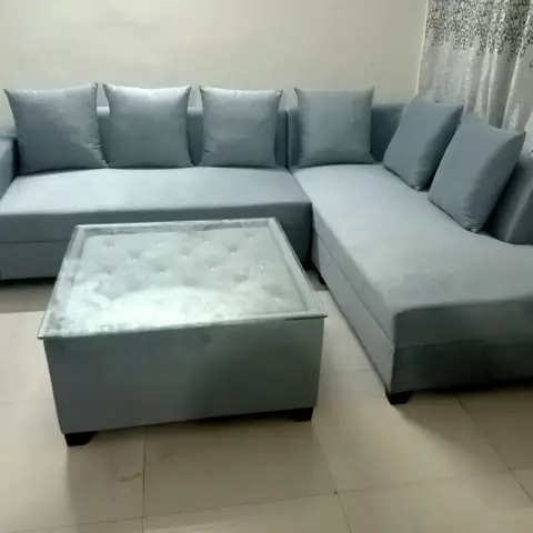 L-shaped grey velvet sectional sofa with a tufted glass-top coffee table against a plain wall with patterned curtains.