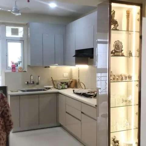 L-shaped kitchen with grey wall units over beige base cabinets, closed off from the dining side by a full-height glass crockery cabinet lit strip-by-strip