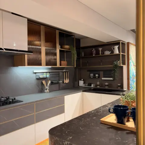 L-shaped modular kitchen in ivory and grey stone-finish handleless shutters with gold gola profiles, fluted amber glass wall cabinets over the hob