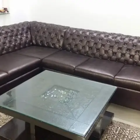 L-shaped tufted brown leather chesterfield-style sofa with a mirrored glass-top coffee table on a patterned rug, styled shelf with decor pieces behind.