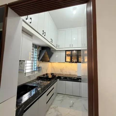 L-shaped white gloss kitchen framed through a fluted wood doorway, with a black marble countertop and backsplash and a glass-front crockery cabinet.