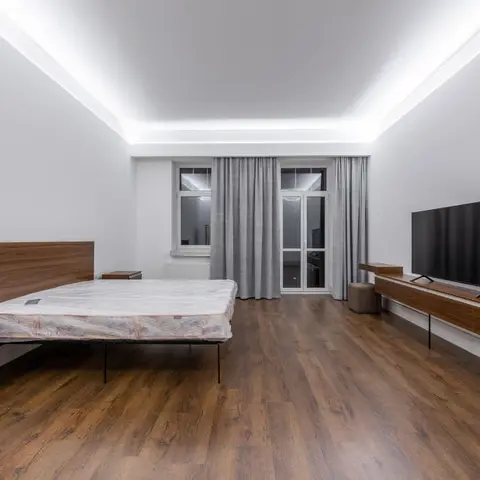 Large bedroom with a bare-mattress bed facing a long walnut floating TV console, cove-lit ceiling and grey curtains at a balcony door.