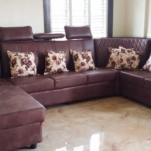 Large brown leatherette L-shaped sectional sofa with floral cushions placed in an otherwise bare living room with marble flooring.