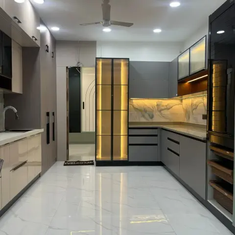 Large L-shaped grey handleless kitchen with a backlit marble-effect splashback, a freestanding black-framed reeded-glass display column lit from inside