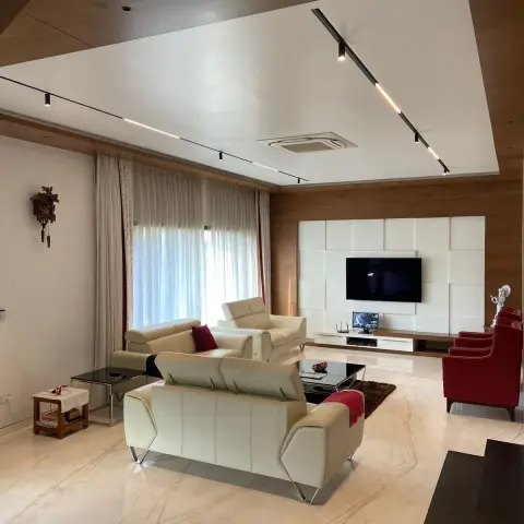 Large living room on cream veined marble floors with cream leather recliner sofas facing a TV wall of white upholstered square pads set into wood veneer