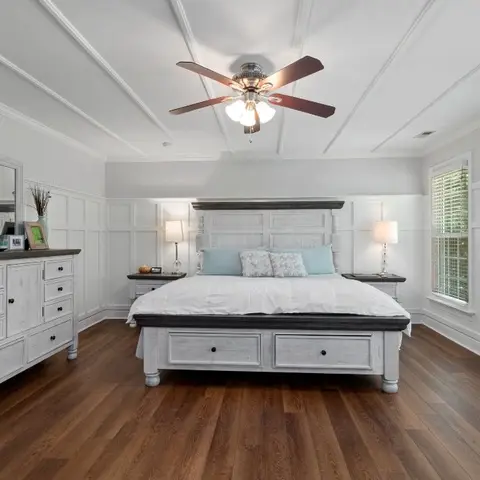 Large primary bedroom with a whitewashed farmhouse-style king bed with drawer storage, a matching dresser with mirror, and panelled wainscoting walls.