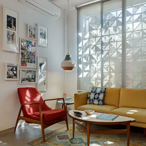 Late afternoon sun striping a mustard three-seater and a red-cushioned teak armchair over a blue-and-ochre kilim
