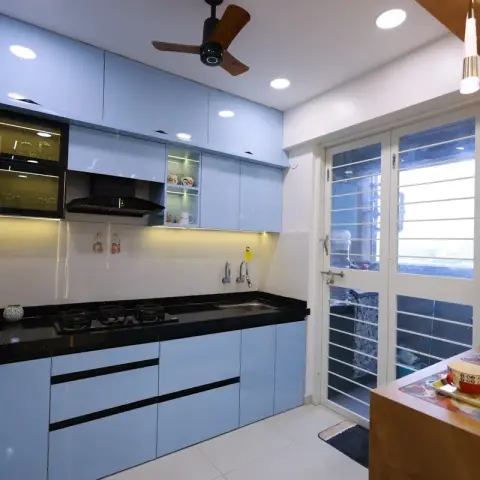 Light-blue glossy modular kitchen with black granite counters, glass-shutter upper cabinets and a lit pooja niche with idols beside the balcony door.