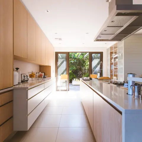 Light-oak galley kitchen with a long quartz-topped island, glass-front upper cabinet, and full-height doors opening onto a garden deck.