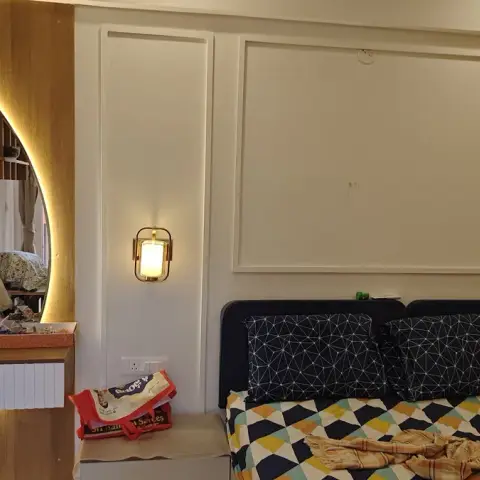 Lived-in bedroom where a crescent-shaped LED-backlit mirror sits over a fluted wall-hung dressing ledge crowded with cosmetics and a red Hanuman idol