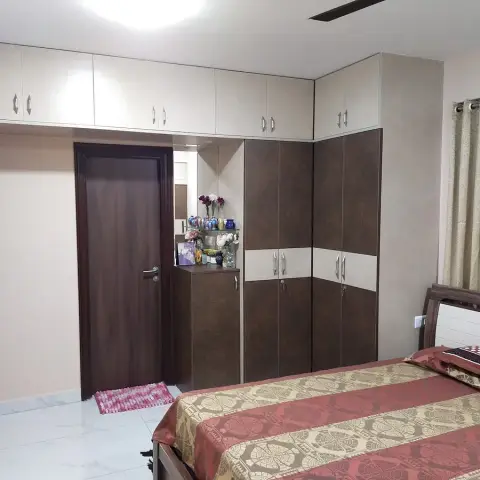 Lived-in bedroom where a two-tone brown-and-cream laminate wardrobe with loft shutters wraps into a corner dressing unit topped with glass shelves of coloured