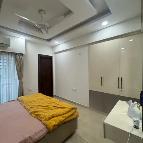 Lived-in bedroom with a rumpled yellow quilt on a low upholstered storage bed, a cream-and-grey high-gloss wardrobe run opposite and a stepped POP ceiling