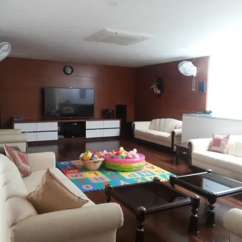 Lived-in family living room with three cream sofas around two carved dark-wood glass-top tables, a teak-panelled TV wall over a white-and-wood console