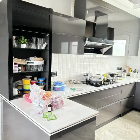 Lived-in grey high-gloss kitchen with a white quartz peninsula, black roller-shutter crockery cabinet standing open on tumblers and dabbas