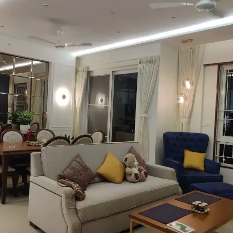 Lived-in living room at night: a beige three-seater with a mustard cushion and a teddy bear faces a navy tufted wingback