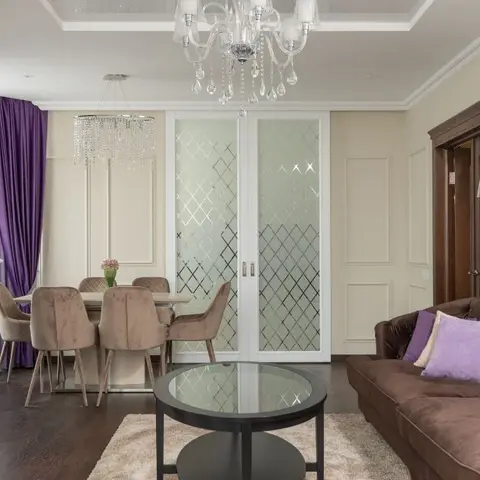 Living and dining space with a crystal chandelier, purple velvet drapes and a brown tufted sofa with lilac cushions beside a diamond-lattice glass door.