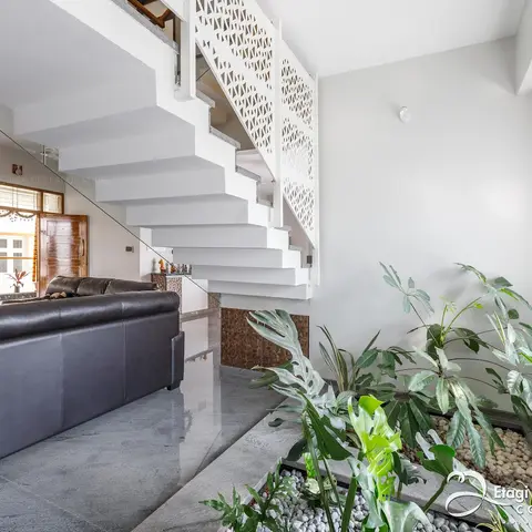 Living area beneath a floating staircase with a white jaali-pattern balustrade, beside an indoor pebble garden and a black leather sofa.