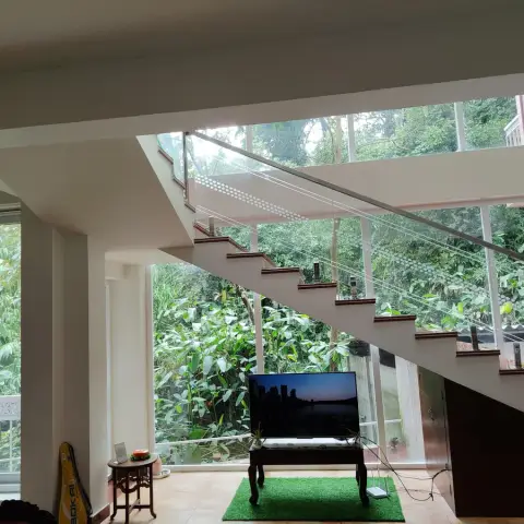 Living area beneath a glass-balustrade floating staircase, with a small wood TV console on an artificial-turf rug and floor-to-ceiling glazing overlooking