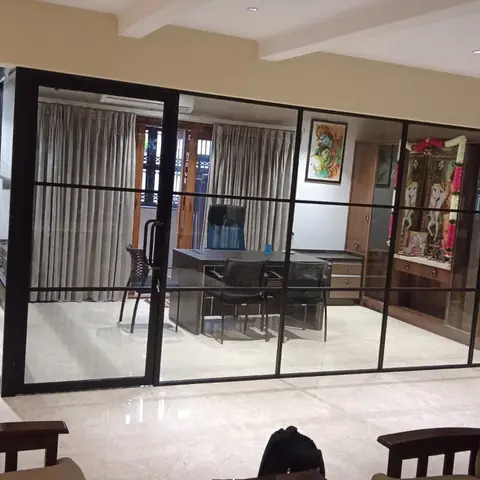 Living area opening through a black steel-framed glass partition into a home office with a desk and chairs.