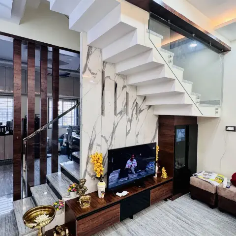 Living area tucked under a white cantilevered staircase with a glass balustrade, TV on a walnut low unit against a book-matched white-and-grey marble slab