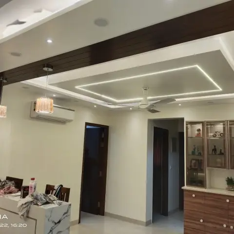 Living area with a rhombus-shaped LED profile cut into the false ceiling, a dark wood ceiling beam, and a glass-front lit crockery display unit