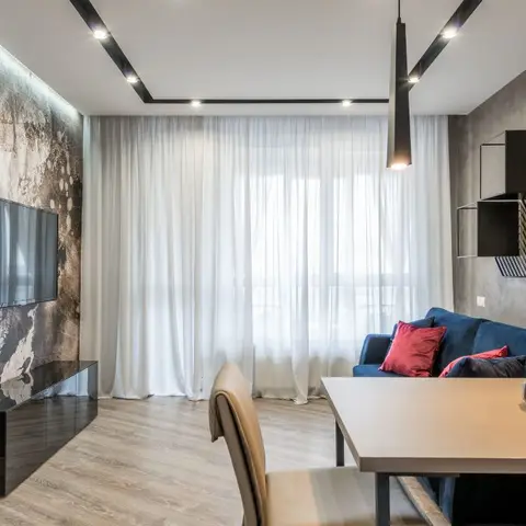 Living-dining space with a textured stone-effect TV wall, slim black pendant lights over the dining table, and a navy sofa with red accent cushions.