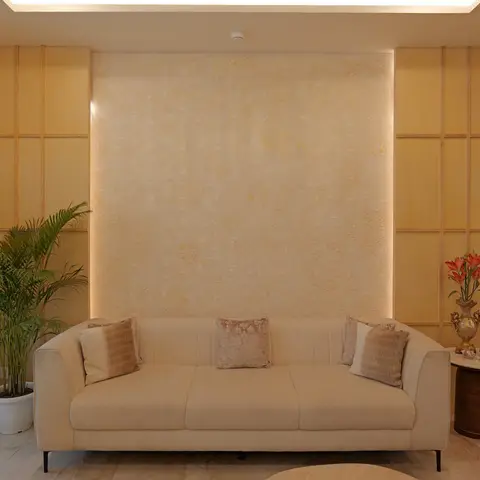 Living room centred on a backlit textured gold-and-cream wallpaper panel flanked by wood panelling, with a cream channel-tufted sofa and a potted areca palm.