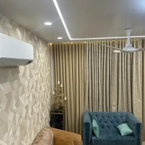 Living room corner where a beige geometric-print wallpaper wall meets full-height champagne curtains lit from a concealed pelmet cove