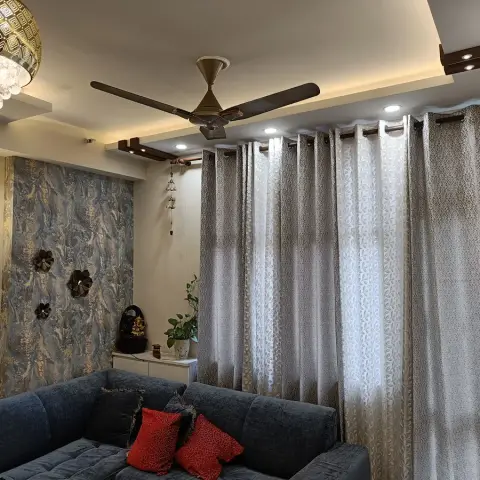 Living room corner where a blue chenille L-sofa with red silk cushions sits under a marbled teal wallpaper panel hung with carved wooden flowers