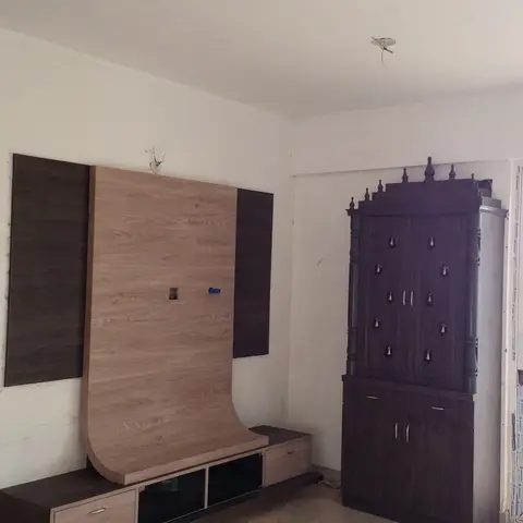 Living room corner with a curved light-wood-veneer TV unit beside a traditional dark-wood carved pooja cabinet