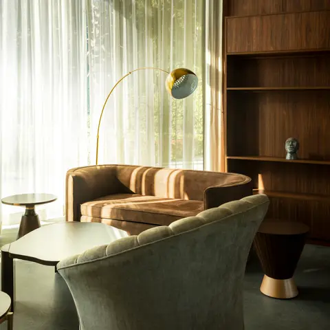 Living room corner with a curved tan velvet sofa, sage tufted armchair, brass arc floor lamp, and a walnut display shelf styled with a sculptural bust.