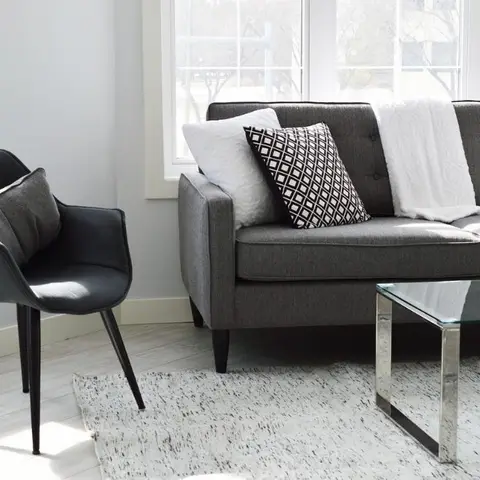 Living room corner with a grey tufted-button sofa piled with mixed-pattern cushions, a black leather accent chair, and a glass-top chrome coffee table