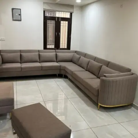 Living room corner with a large taupe L-shaped sofa on gold legs and matching ottomans, a jaali-cut room divider beside the window.