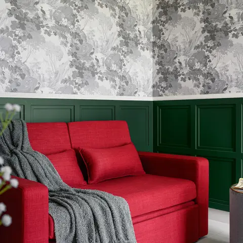 Living room corner with a red upholstered sofa and grey throw against dark green wainscoting paneling topped by a grey toile-print wallpaper with birds