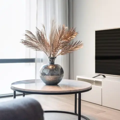 Living room corner with a round marble-top coffee table holding a bronze vase of dried palm fronds, a white low TV console, and sheer curtains behind.