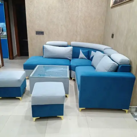 Living room corner with a two-tone teal and light-grey L-shaped sofa, matching upholstered ottomans, a glass-top coffee table