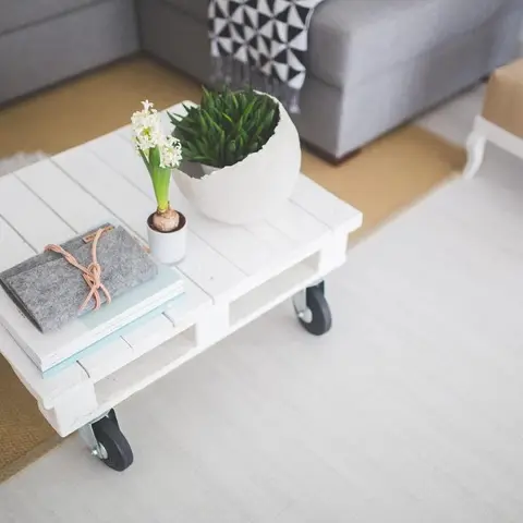 Living room corner with a white pallet-style coffee table on casters, layered fur and jute rugs, and a grey sofa with a black-and-white triangle-print throw.