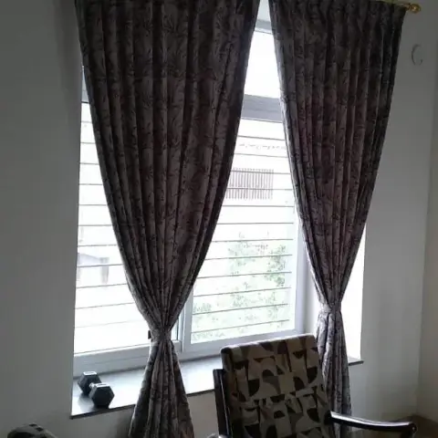 Living room corner with floral-patterned floor-length curtains on a brass pole, a carved wood armchair and sofa beside a grilled window.