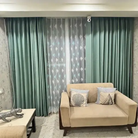 Living room corner with teal pinch-pleat drapes over leaf-embroidered sheers, a beige two-seater carrying shell and sailboat cushions